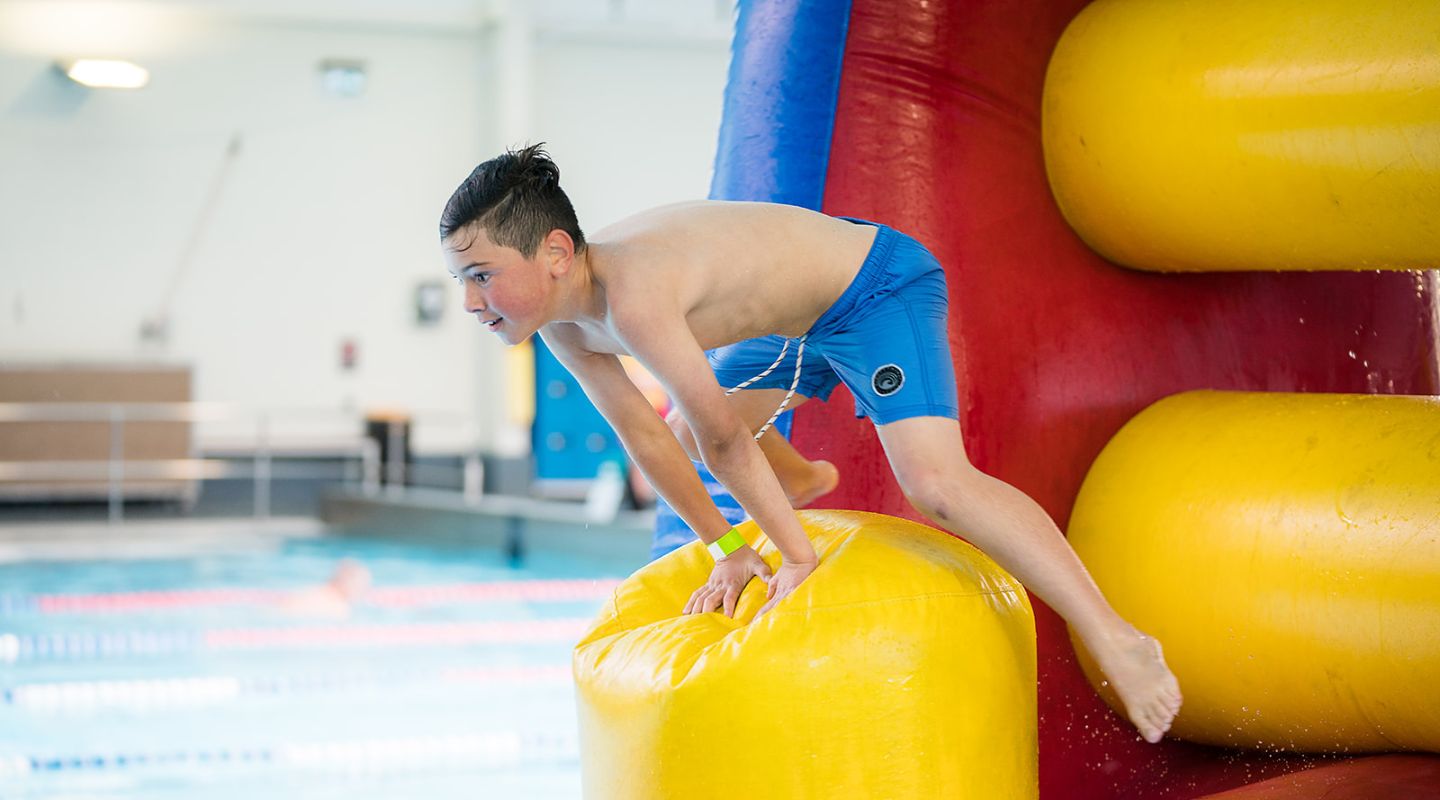 A boy jumping off of a yellow inflatable obstacle course into the Hornby Rec and Sport Centre pool. 