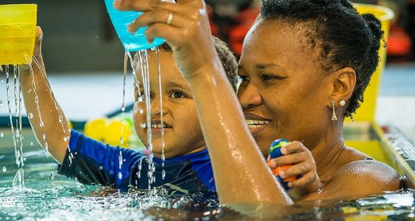 A little boy holding a yellow cup while being held by a smiling woman at Christchurch Recreation and Sport pool. 