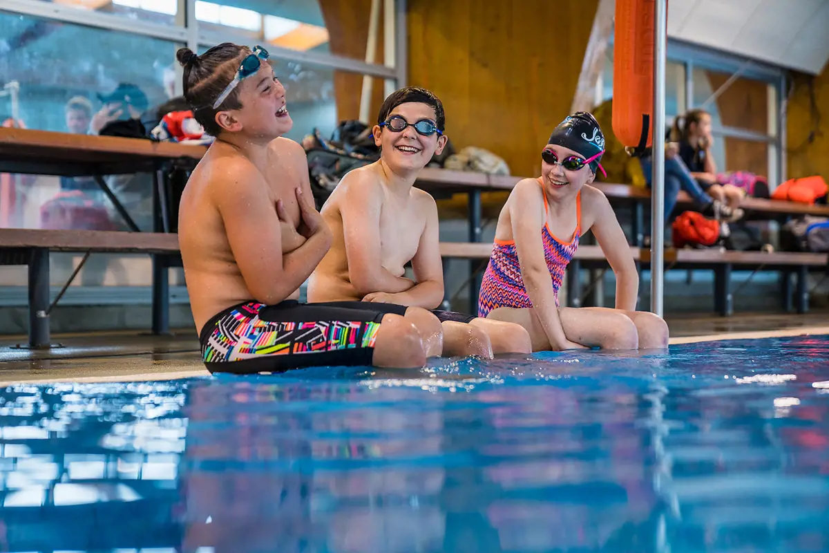 A group of smiling children sitting on the edge of the pool deck at Christchurch Recreation and Sport pool.