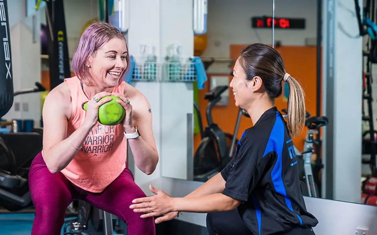 A female personal trainer assisting a woman in pink through a kettle bell workout at Christchurch Recreation and Sport Centre.