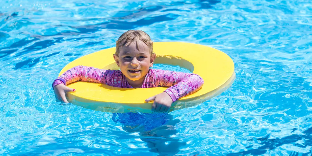 A smiling girl in pink swimmers holding onto a yellow floating ring at Waltham Summer pool.