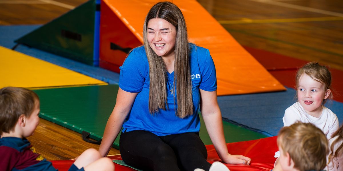 A group of children and a smiling instructor sitting on mats during a pre-school gymnastics class at Christchurch Recreation and Sport centre. 