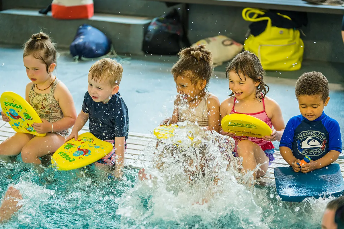 A group of five smiling children sitting on the pool edge splashing at Pioneer Recreation and Sport Centre.