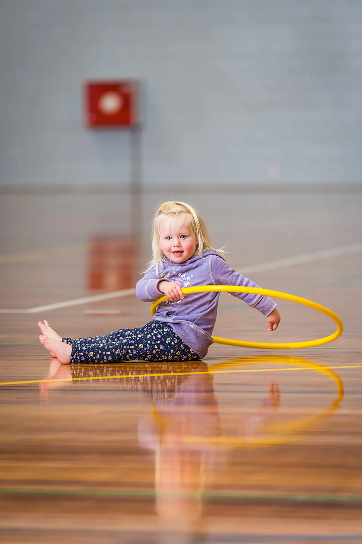 A smiling toddler playing with an orange hula-hoop during a Tumbletimes class at Christchurch Recreation and Sport Centre.