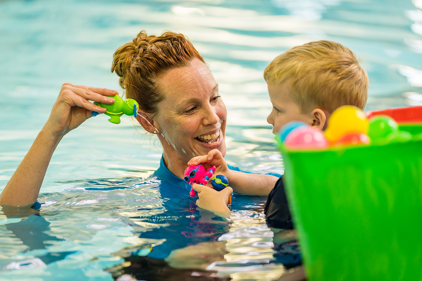 A smiling swim instructor holding a little boy who is playing with pool toys at Christchurch Recreation and Sport pool. 