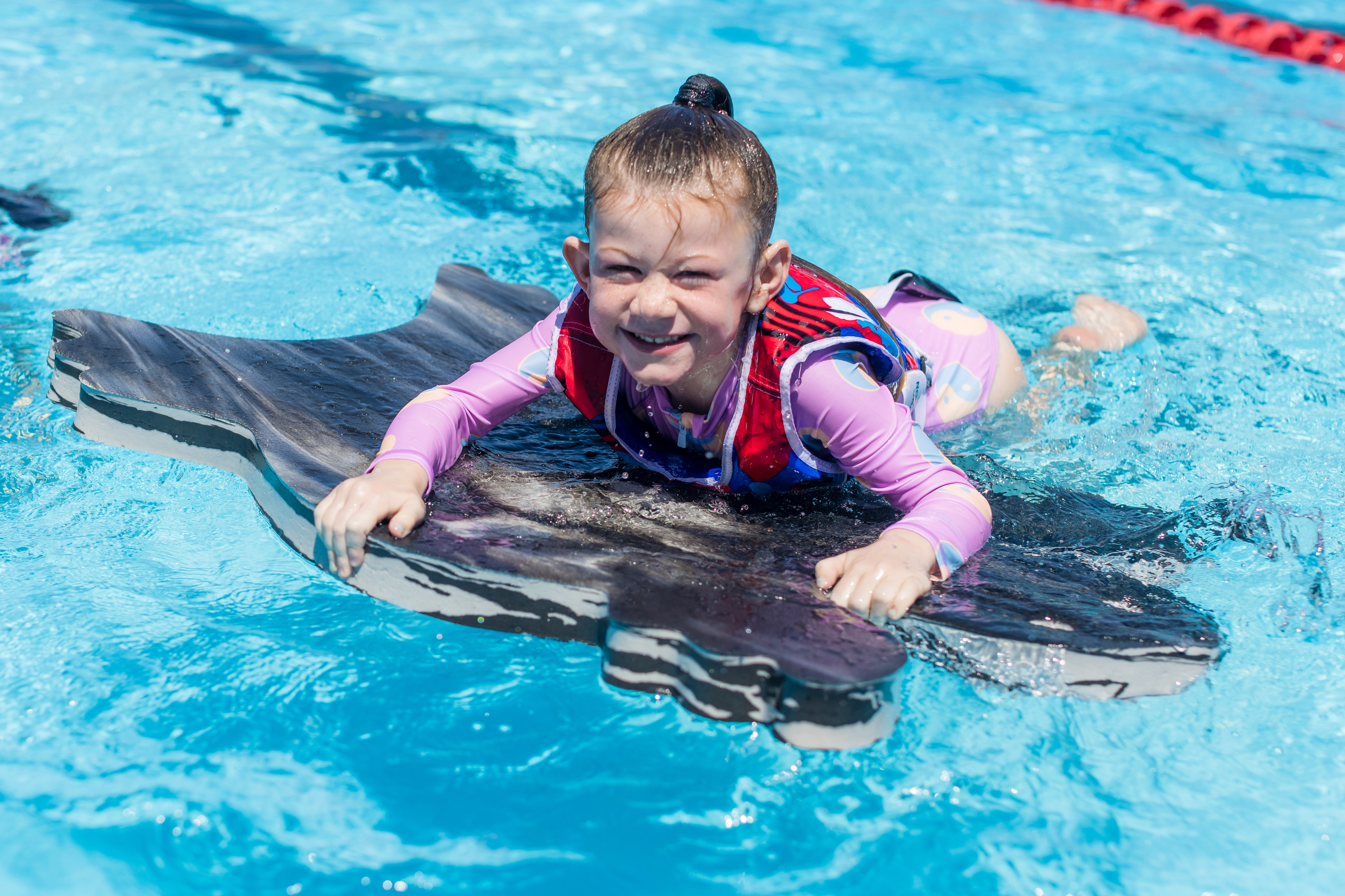 A smiling girl wearing pink holding onto a black flotation mat at the Halswell Summer pool. 
