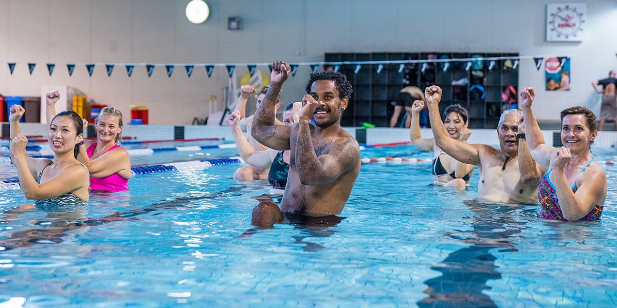 A group of eight smiling adults participating in an Aqua Zumba class with their arms above their heads at Christchurch Recreation and Sport pool.