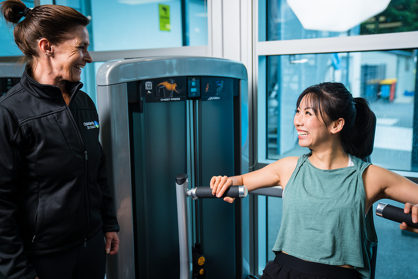 A female personal trainer in a black staff uniform assisting a female client with a shoulder press at Christchurch Recreation and Sport Centre.