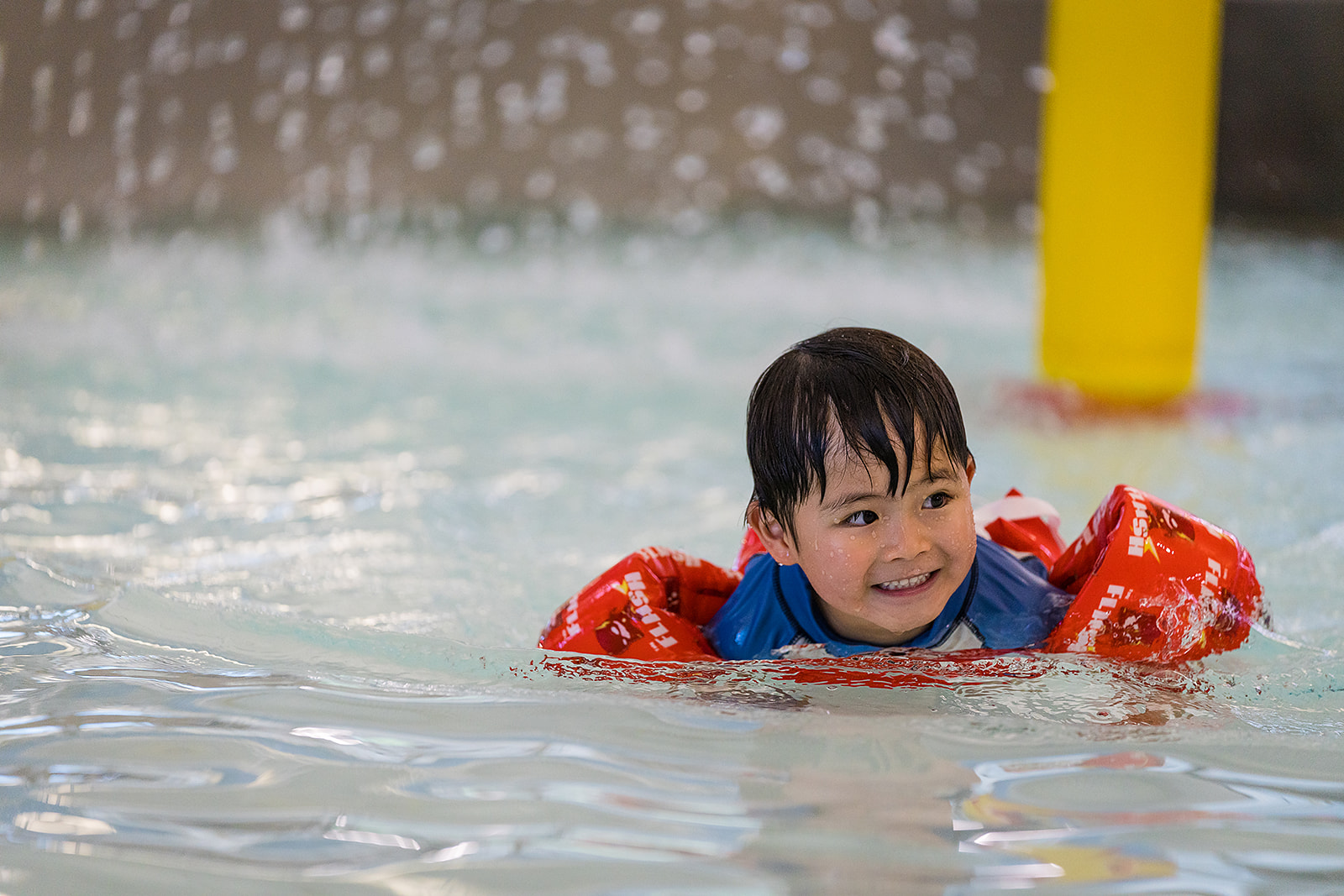 A smiling boy wearing a red lifejacket swimming at Christchurch Recreation and Sport pool. 