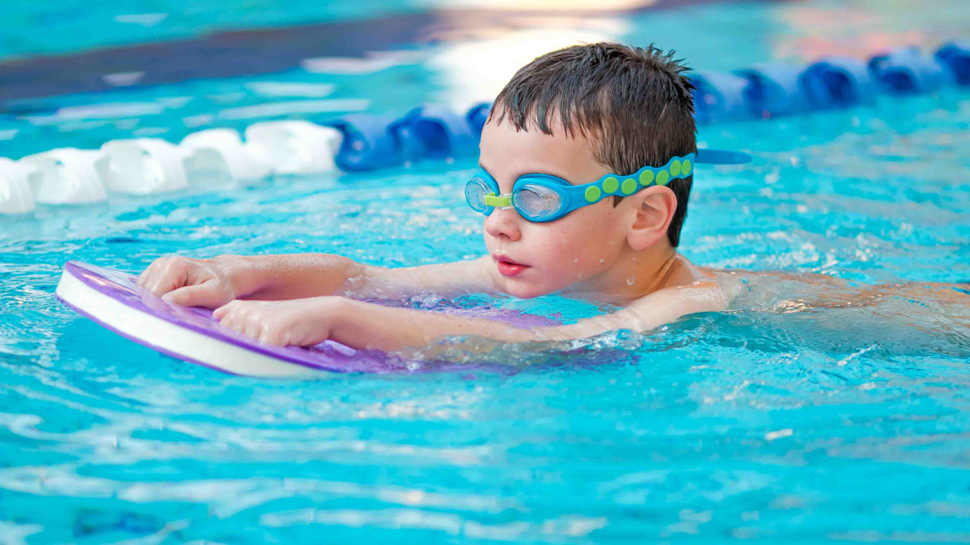 A little boy in goggles swimming with a purple flotation board at Christchurch Recreation and Sport pool. 