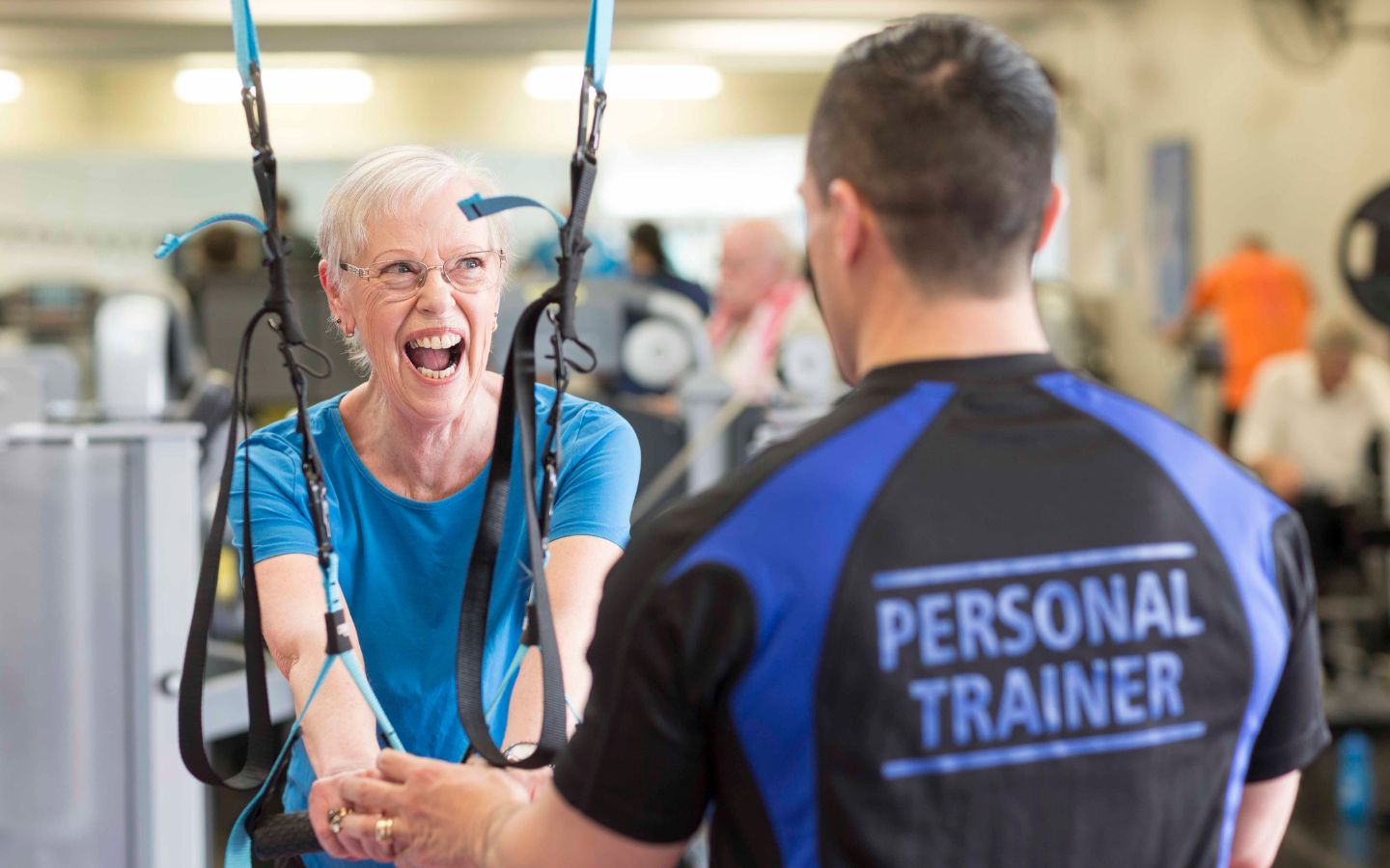 A male personal trainer in a blue and black shirt guiding a smiling elderly woman through an arm workout at Christchurch Recreation and Sport centre. 