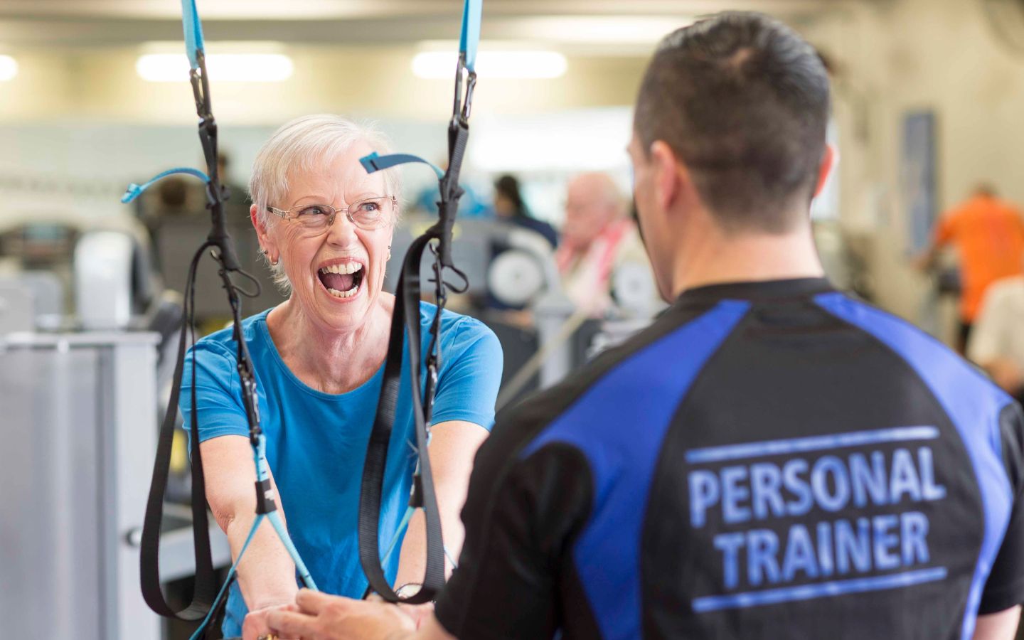 A male personal trainer in a blue and black shirt guiding a smiling elderly woman through an arm workout.