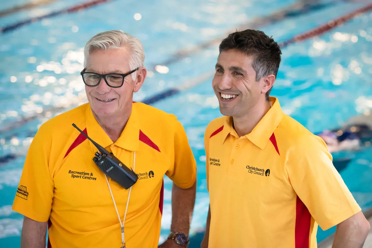 Two smiling male lifeguards in yellow staff uniform standing on the pool deck at Christchurch Recreation and Sport pool.