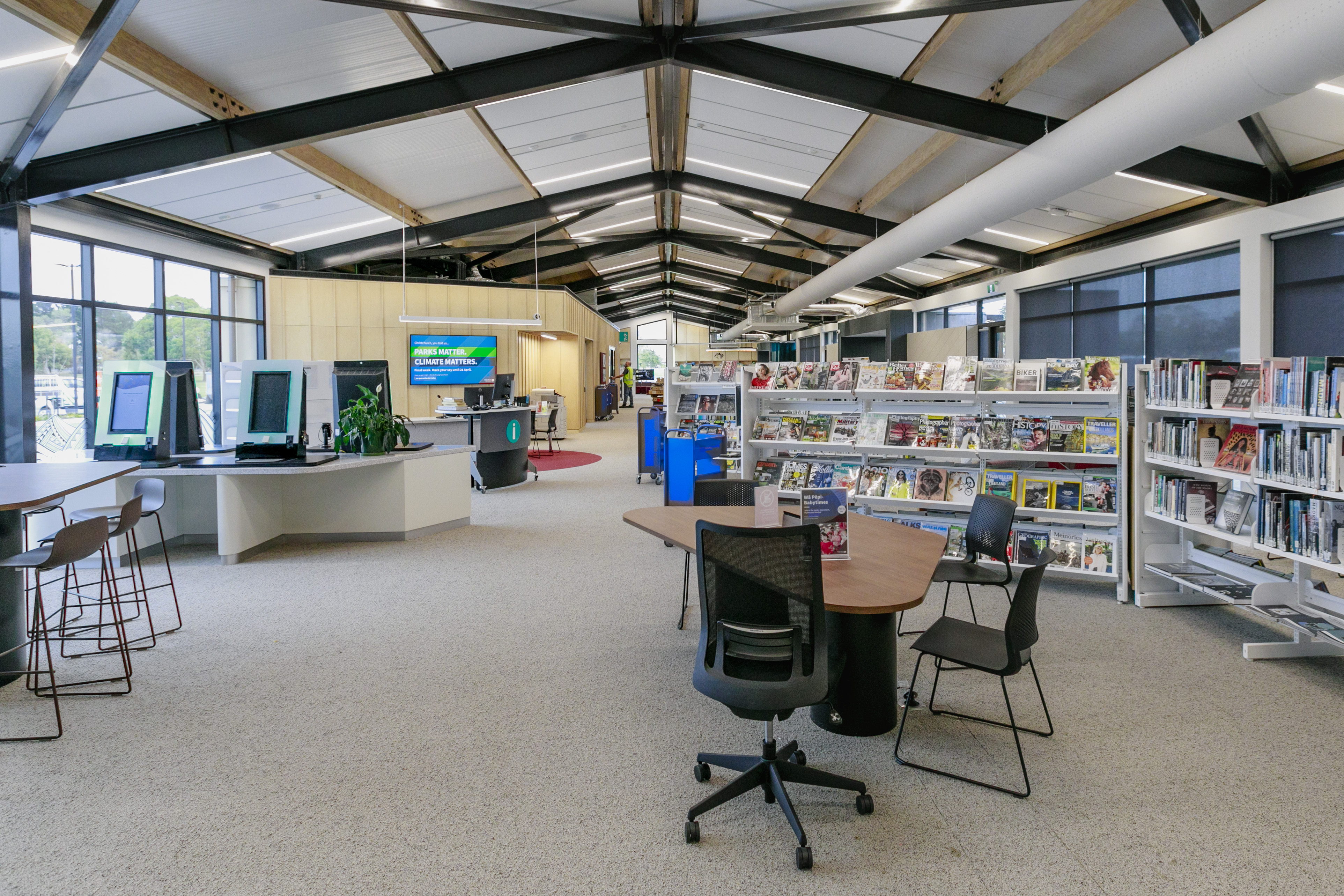 The Matatiki Hornby library equipped with white shelves of books, a large wooden table with black chairs, monitors for book sign-out and an information desk. 