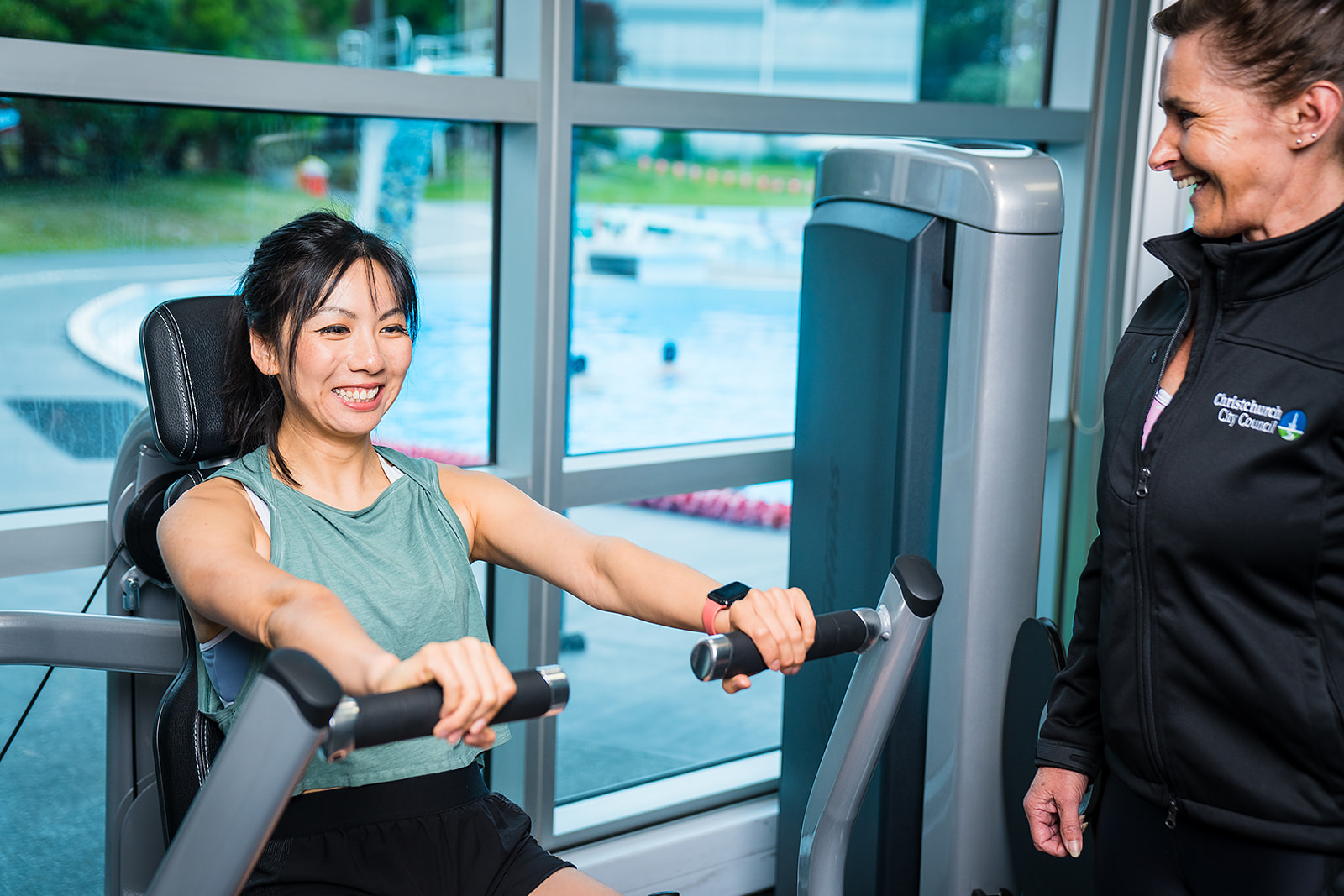 A female personal trainer in a black staff uniform assisting a female client with a shoulder press at Christchurch Recreation and Sport Centre.