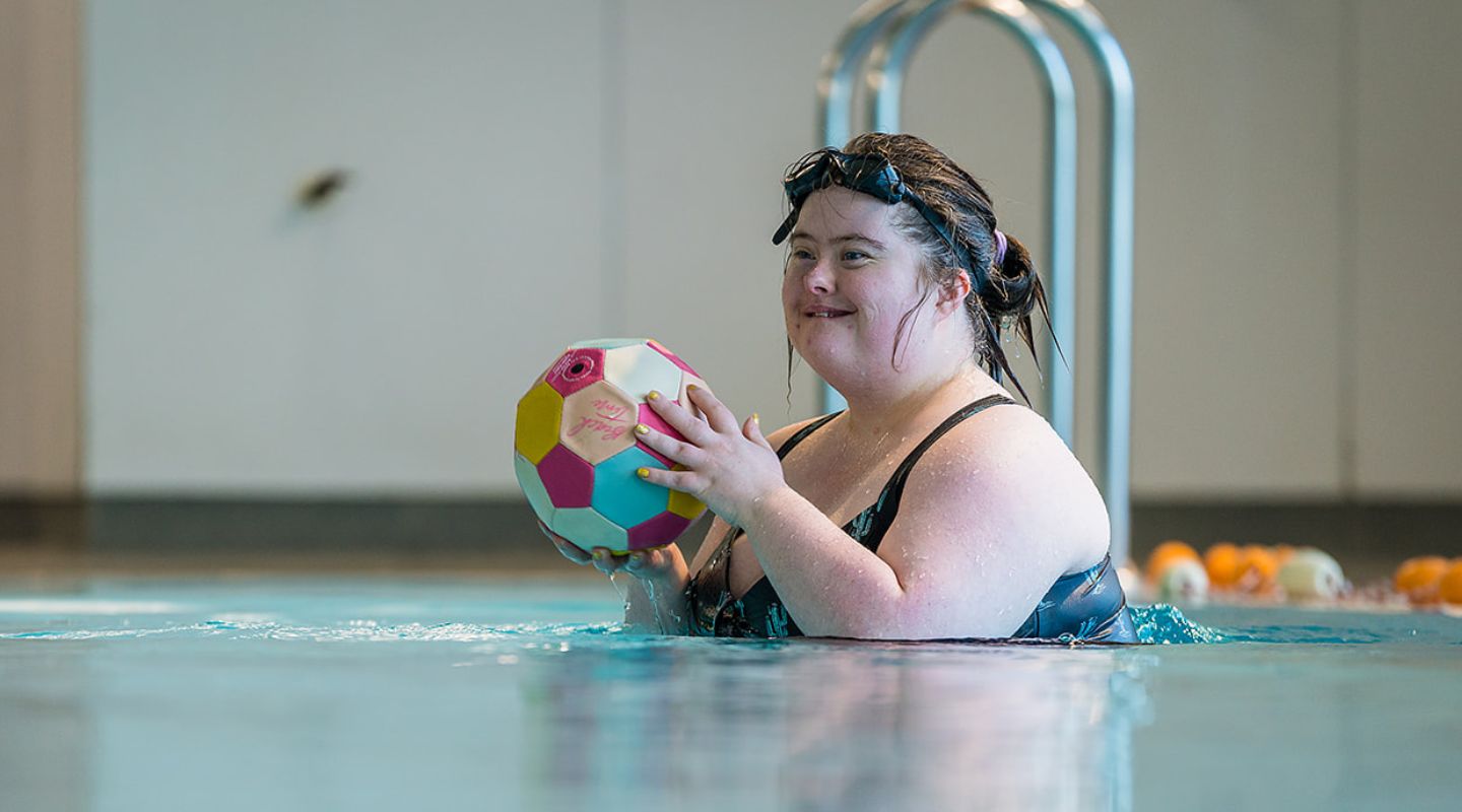 A smiling woman playing in the pool with a yellow and pink football during a Sensory Swimming class at Christchurch Recreation and Sport pool.