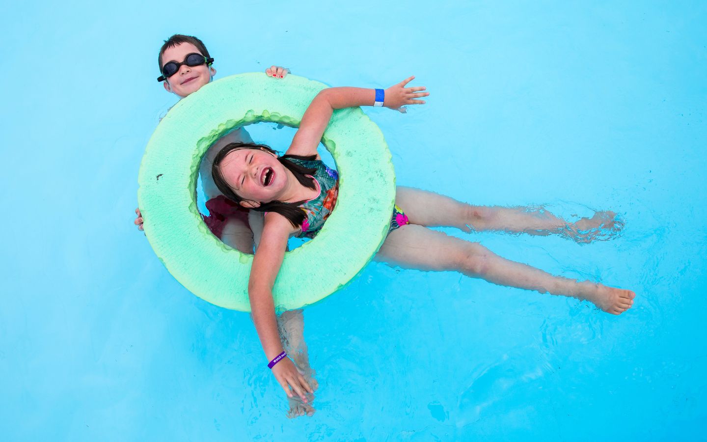 A smiling girl in an inflatable ring floating with a little boy holding onto the side at Templeton Summer Pool. 