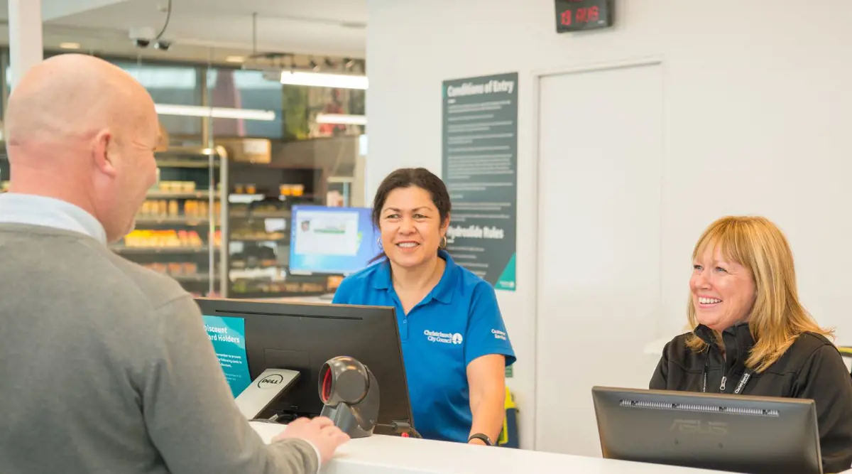 Two smiling women standing behind the counter at Jellie Park Rec Centre talking to a man in a grey sweater.