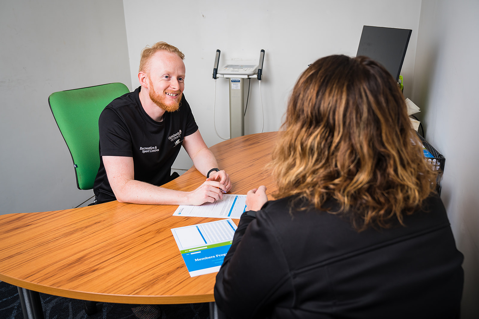A male personal trainer in a black staff uniform sitting across a desk from a female client at Christchurch Recreation and Sport Centre.