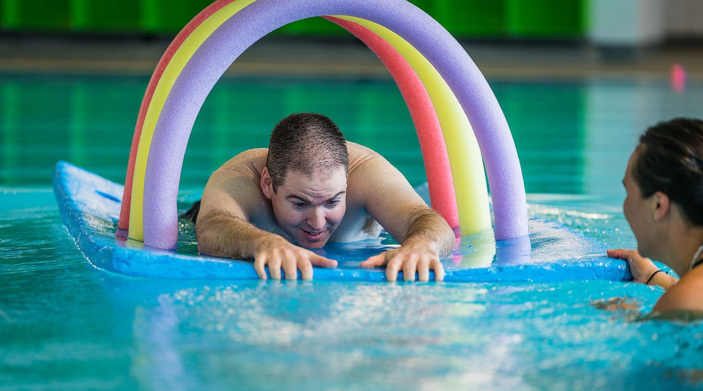 A smiling man playing on a floating blue pool mat during a Sensory Swimming class at Christchurch Recreation and Sport pool.