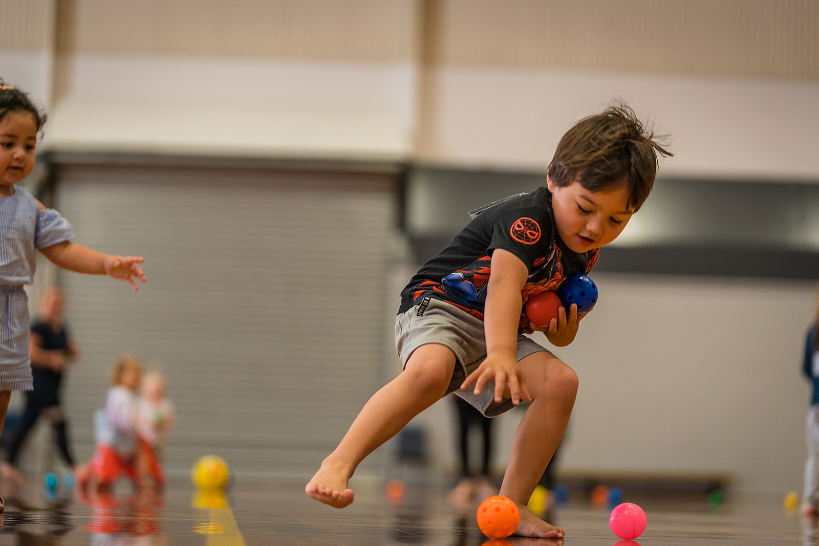 A little boy picking up a handful of brightly coloured balls during a Pre-school sports class at Graham Condon Recreation and Sport Centre. 