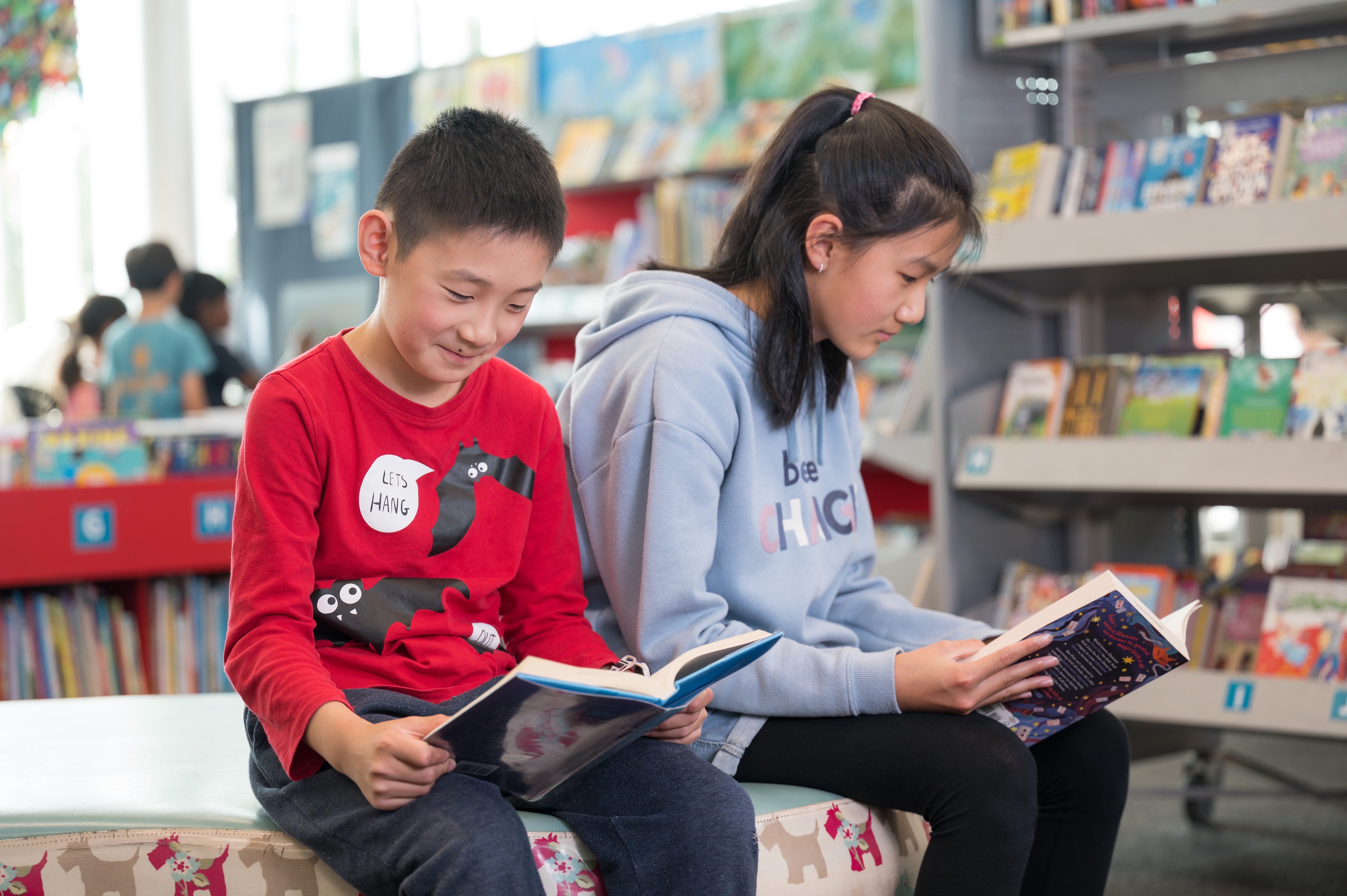 Two children, one younger boy and one older girl seated in the library each reading a book. 