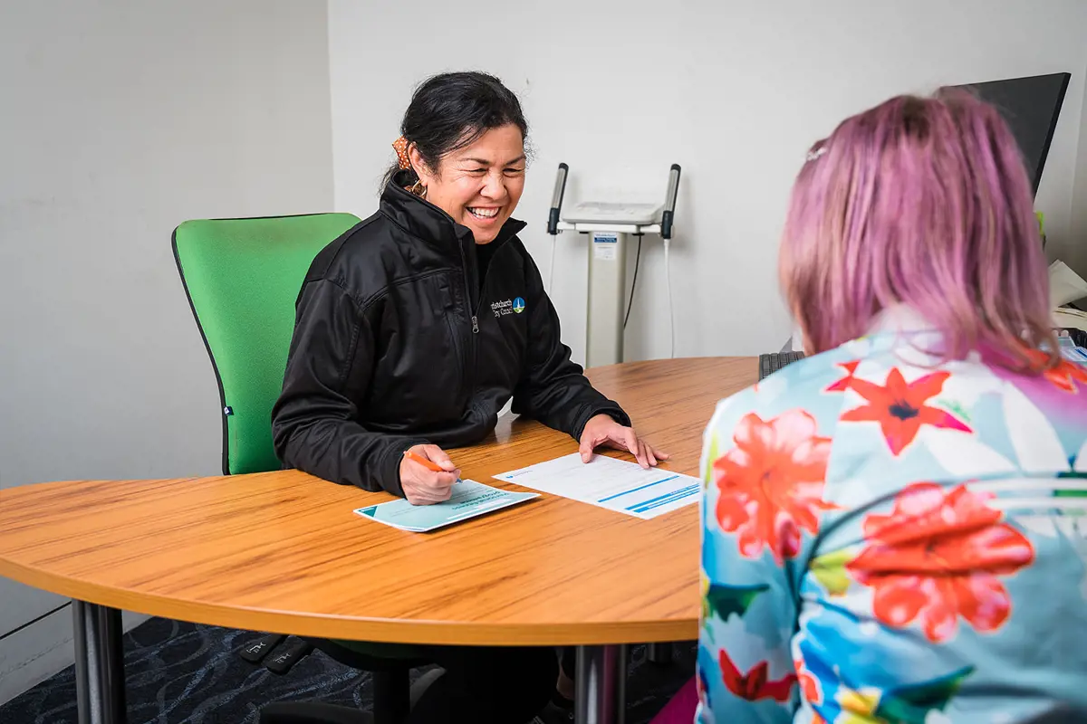 A smiling female personal trainer in a black staff uniform sitting across a desk from a female client at Christchurch Recreation and Sport Centre.
