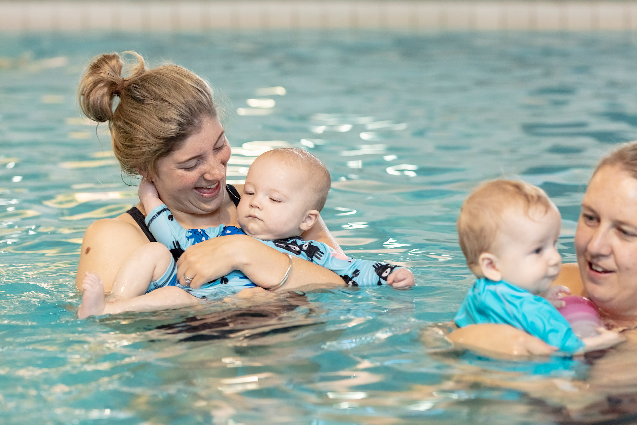 Two smiling women holding onto babies while attending a Tiny Turtles swim class at Pioneer Recreation and Sport Centre. 