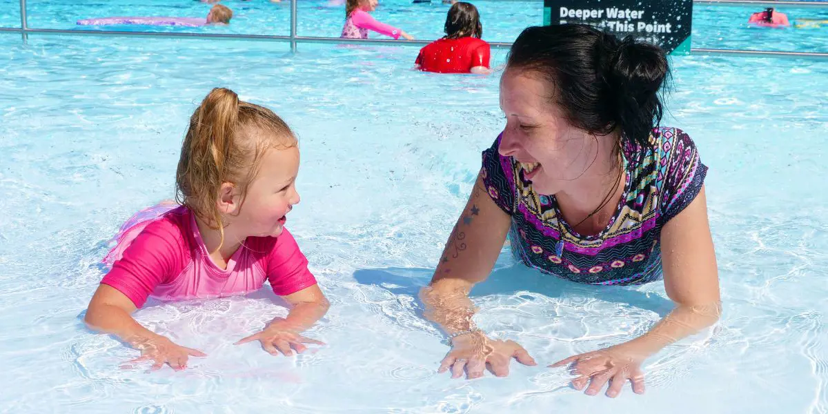 A child in pink and a smiling woman lay on their stomachs in the toddler pool at a Christchurch summer pool.