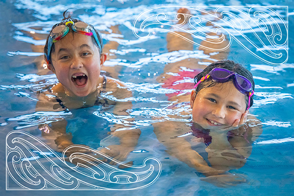 Two young smiling girls with goggles on their heads swimming in the shallow end of Matatiki Hornby Centre pool. 