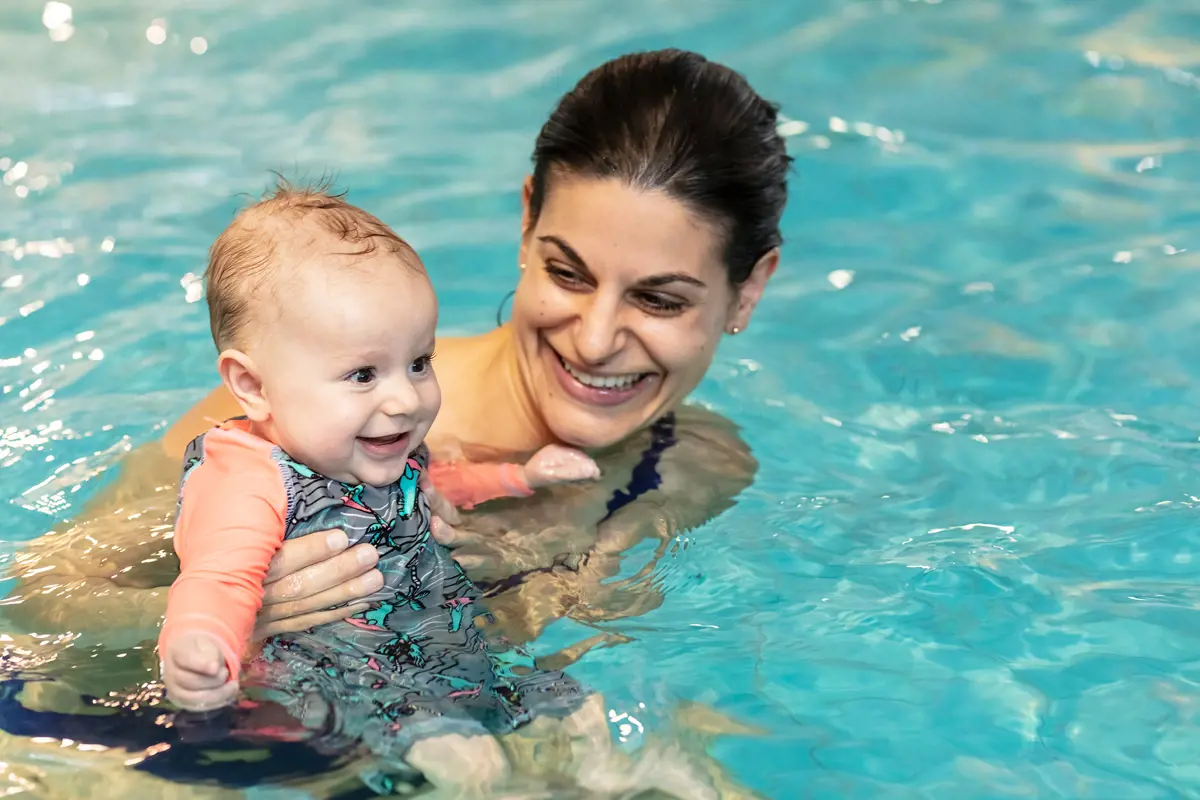 A smiling woman holding onto a baby while attending a Tiny Turtles swim class at Pioneer Recreation and Sport Centre.